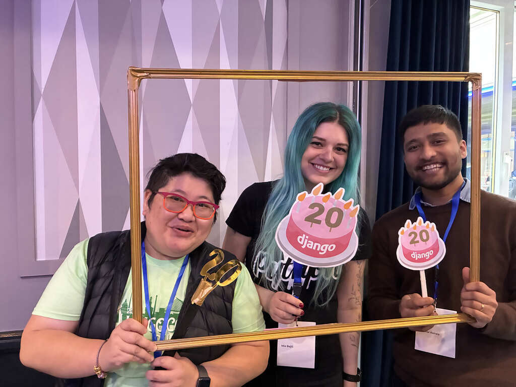 Three people in a group photo, smiling, holding a golden picture frame. Two hold birthday cake props that say 'Django' and '20'
